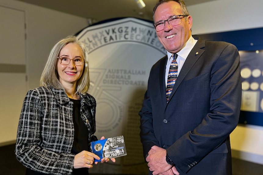 A man and a woman, both in suits, are smiling at the camera as they stand in front of a very large 50 cent coin.