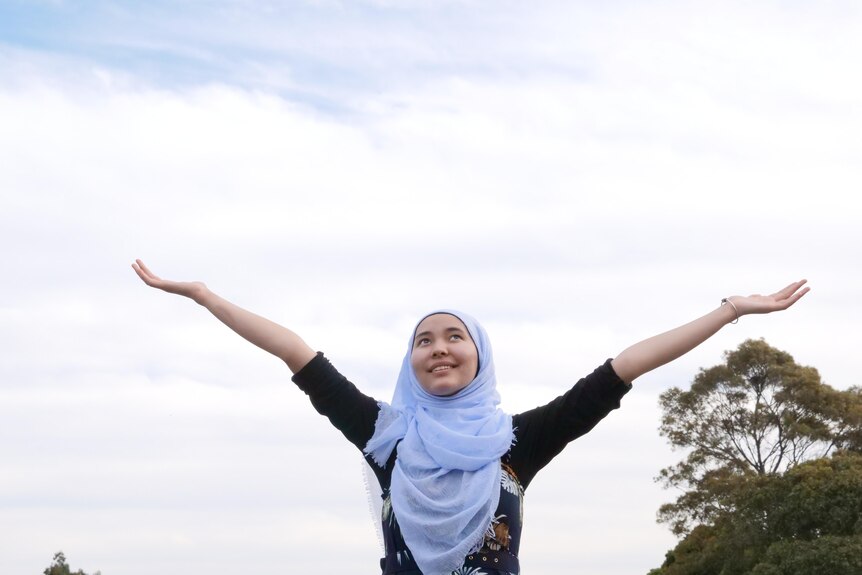 A teenager in a hijab raises both arms to the sky. She looks up with a smile.