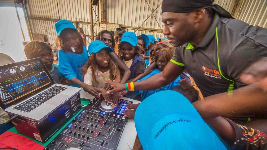 Indigenous Australian children laugh and smile as a man of African appearance teaches them how to use a DJ mixing deck.