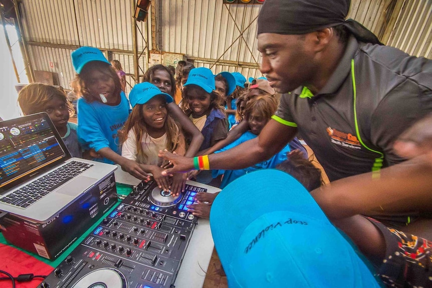 Indigenous Australian children laugh and smile as a man of African appearance teaches them how to use a DJ mixing deck.