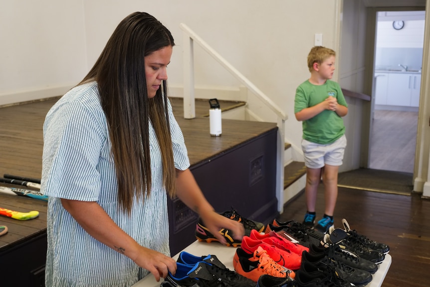 A woman stands behind a table with dozens of football boots on it, a boy in the background.