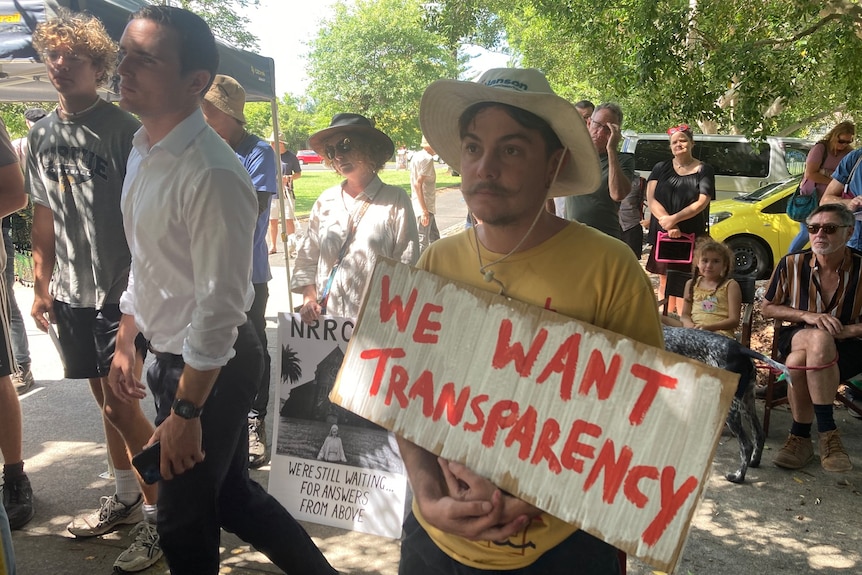 A man in a hat and a yellow tee short holds a sign saying "We want transparency".