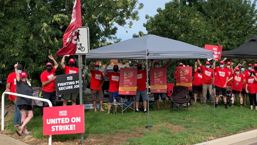 A group of people wearing red t-shirts and masks and holding signs gather on a street, under a gazebo.