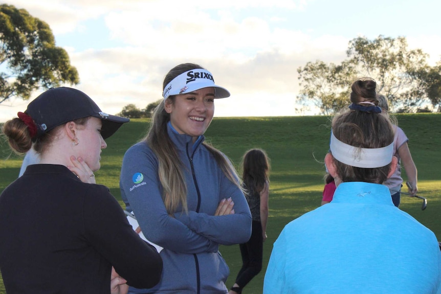 A female golf player in a white cap and grey jumper laughs and talks to children on a golf course.