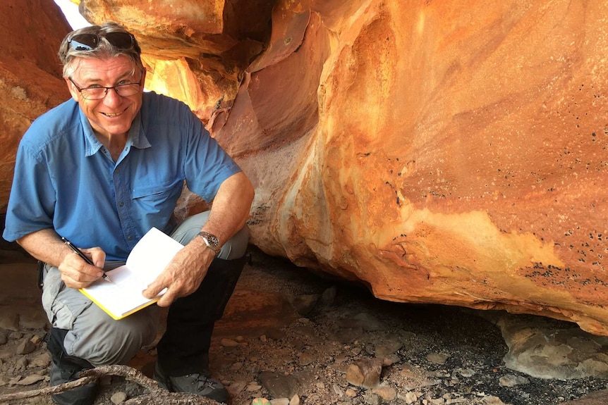 Richard Cosgrove smiles while holding a notebook and crouching near a rock face featuring rock art.