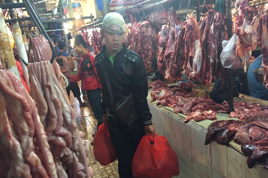A young man carries bags through a market in Indonesia surrounded by hanging meat.