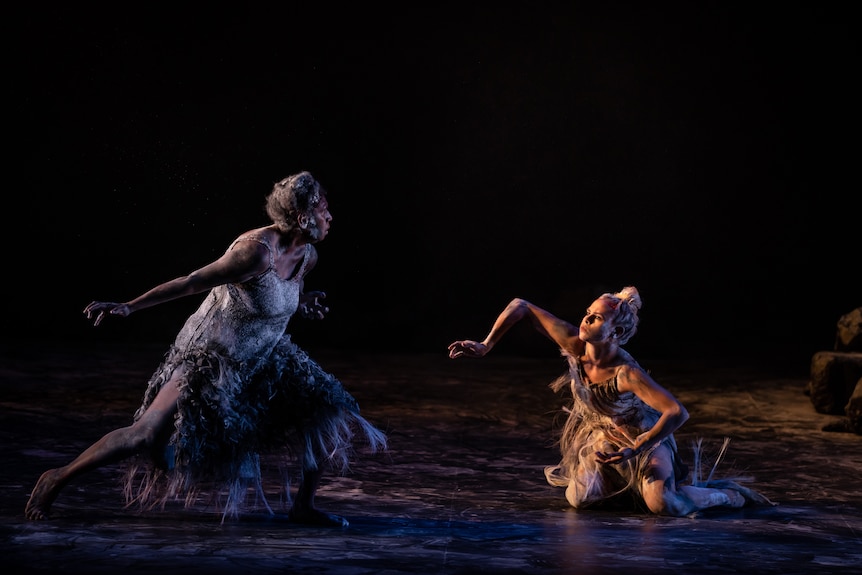 Two Indigenous women performers - one older and one younger - perform together on a dark stage, the younger woman on her knees