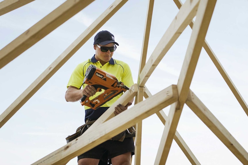 A photo of a carpenter holding a drill on a roof at construction site