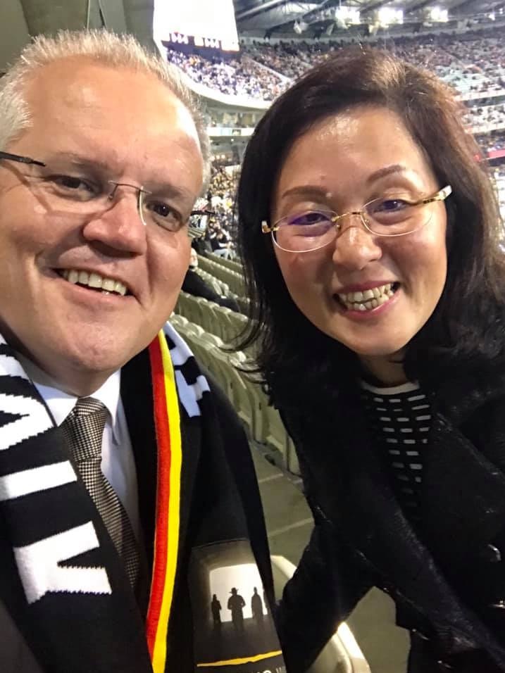 Scott Morrison wearing glasses, suit and scarf next to Gladys Liu, wearing glasses. with football stadium  in background