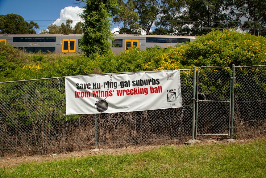 баннер с надписью «Спасите пригород Ку ринггай» висит на металлическом заборе в парке на фоне поезда