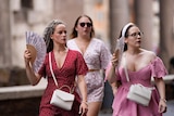 Three women in bright coloured dresses walk along Romes streets with foldable fans