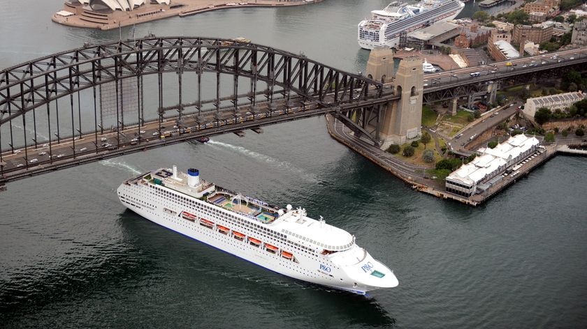 A large cruise ship motors under the Sydney Harbour Bridge and past large waterfront buildings.