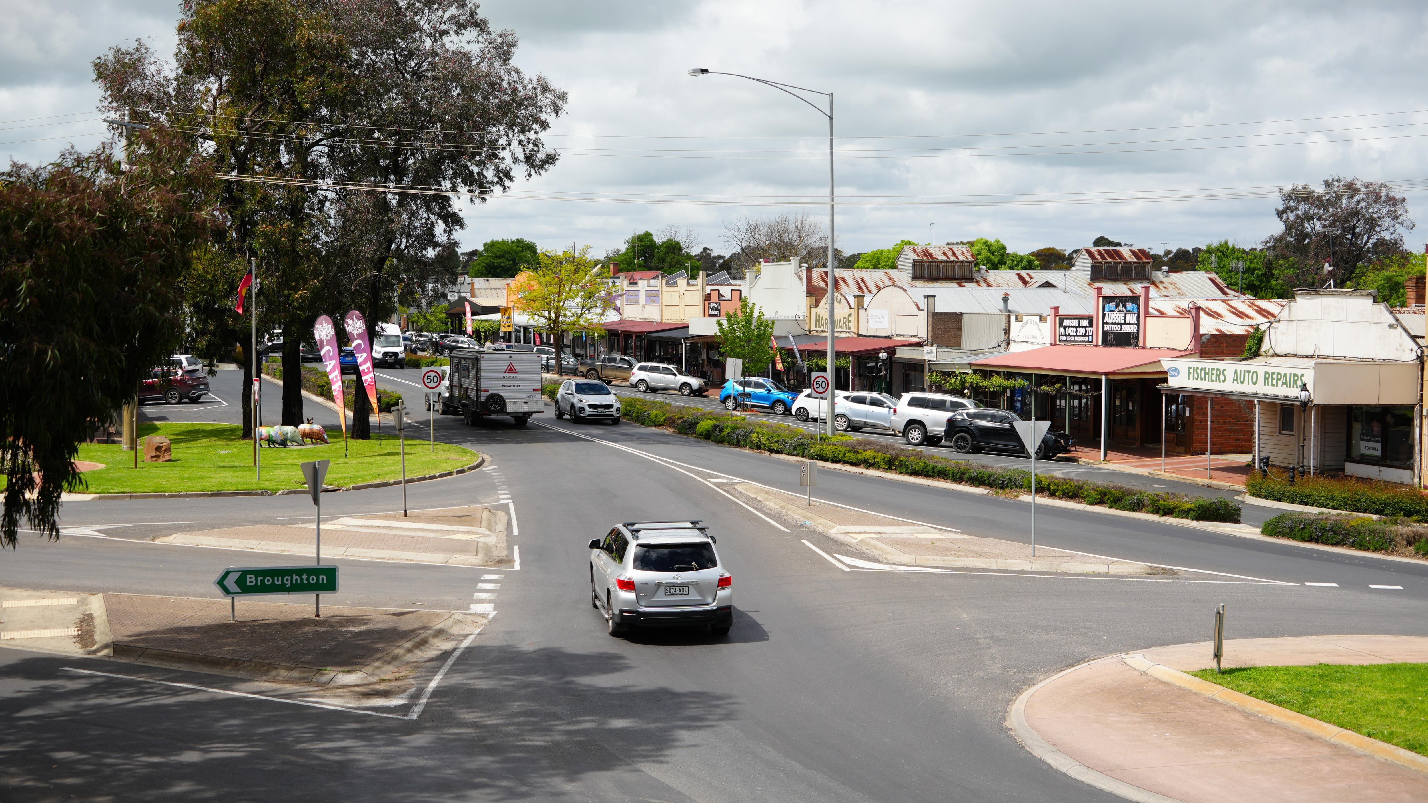 A road through a town, with shops on the right-hand side and a sign pointing left to "Broughton".