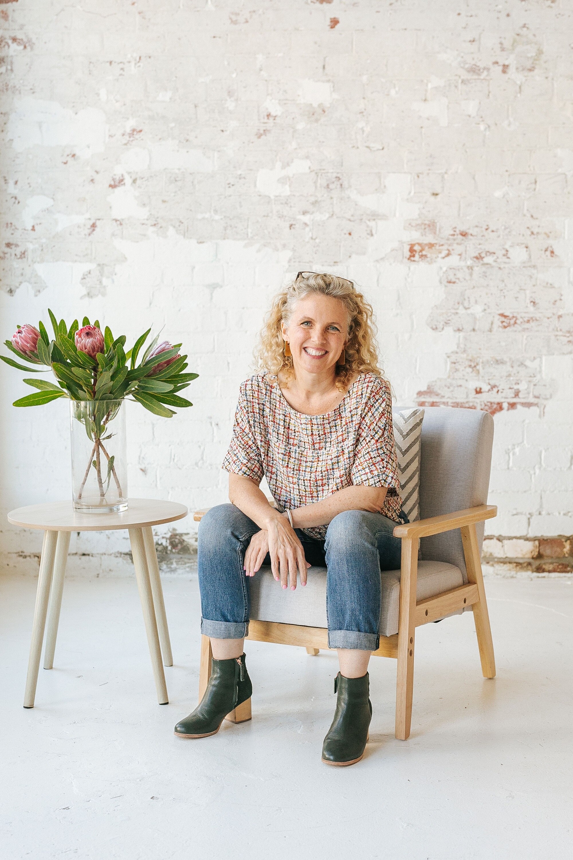 A white woman with curly blonde hair and wearing jeans sits in an armchair beside a table with a vase of flowers on it