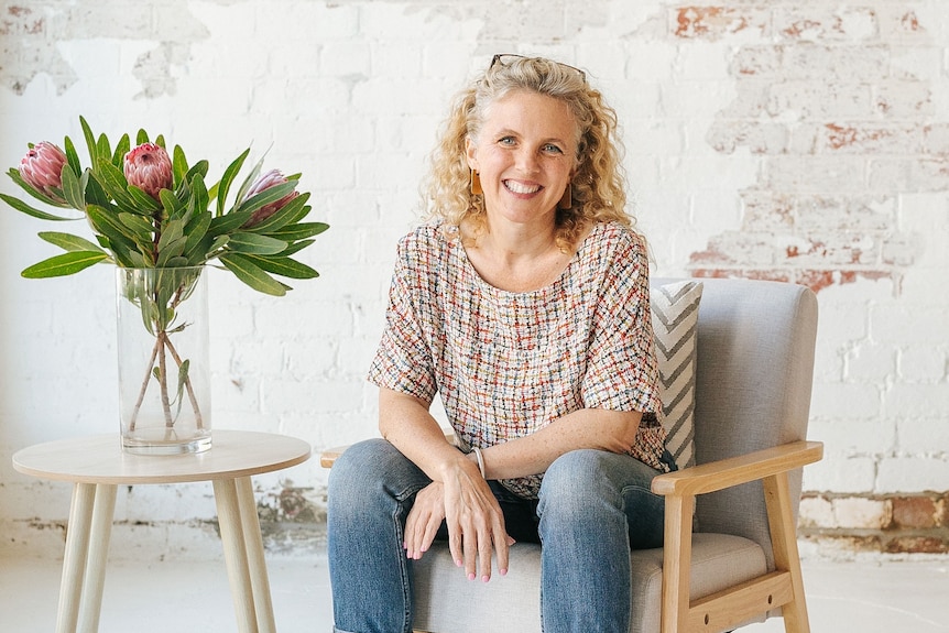A white woman with curly blonde hair and wearing jeans sits in an armchair beside a table with a vase of flowers on it