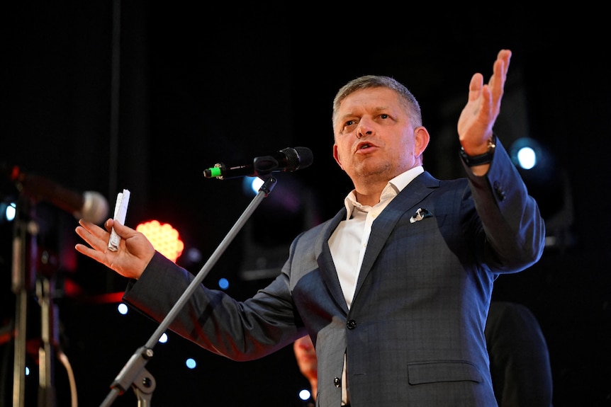 A man stands behind a microphone, speaking and gesturing with his hands.