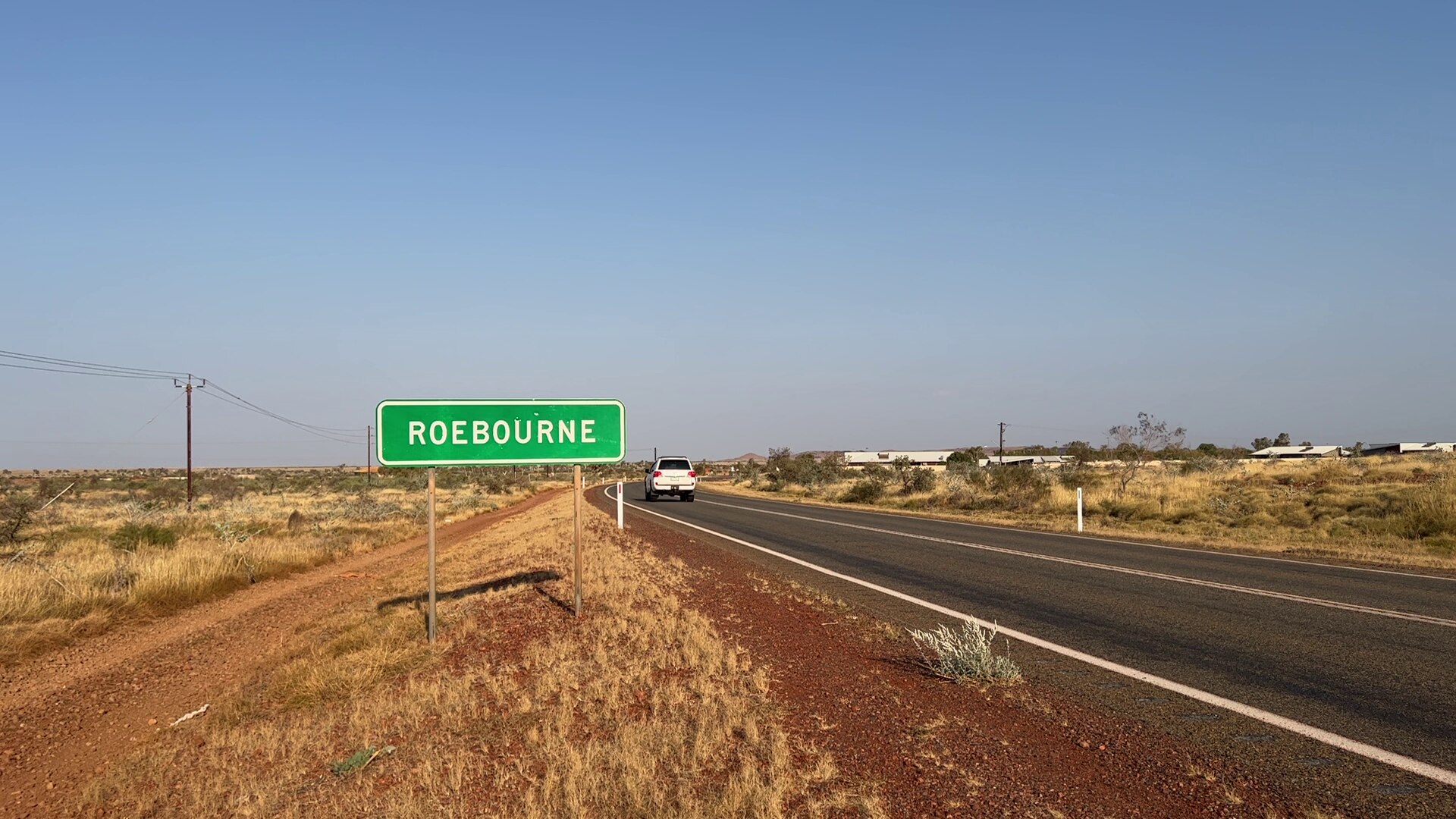 A white four-wheel drive passes a street sign that reads 'Roebourne'.