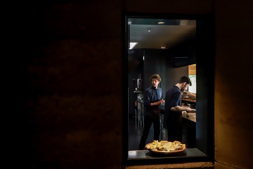 Illuminated window in stone wall looking into a kitchen with two black shirt clad men preparing food