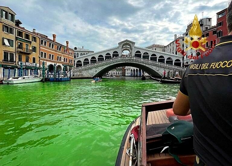 A person on boat on a canal with bright green water, a bridge visible in the background. 