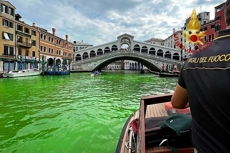A person on boat on a canal with bright green water, a bridge visible in the background. 