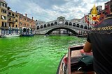 A person on boat on a canal with bright green water, a bridge visible in the background. 