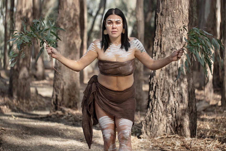 Native woman wearing brown scarf around chest, scarf around waist, paint on face, body, holds shrubs, stands among trees.