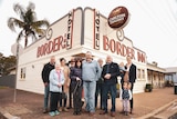 A group of men and women stand on a street corner in front of a large building with the words 'Border Inn'.