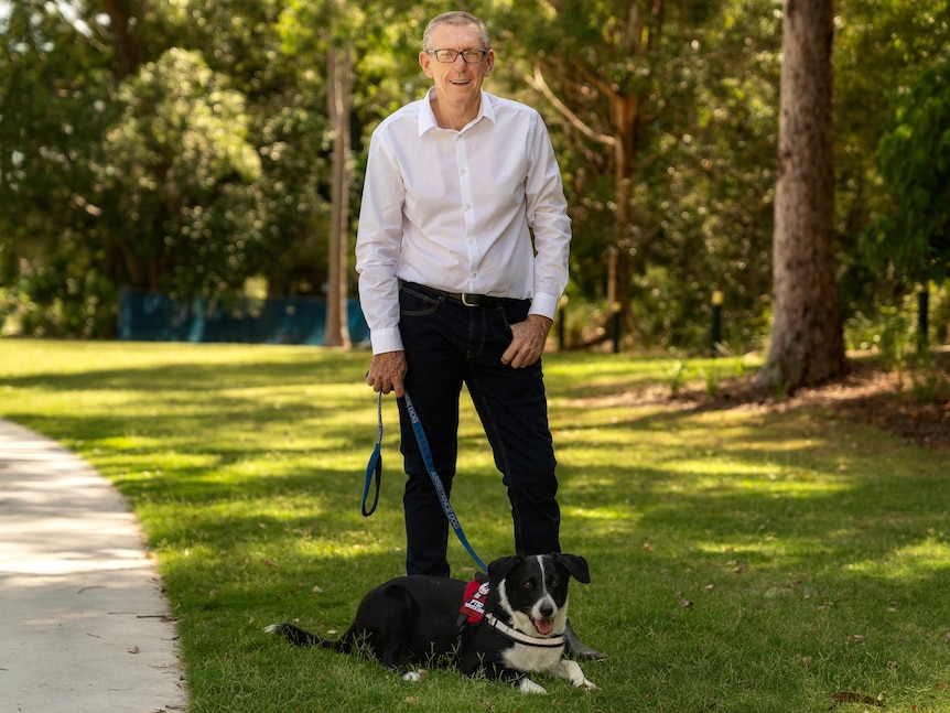 A middle-aged white man with short hair, a white shirt and glasses standing in a park with a border collie