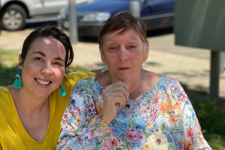 A young woman with brown hair crouches down next to an older woman wearing a floral shirt in a park on a sunny day.