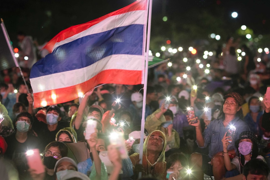 People sitting in rain coats waving lights from smartphones and a Thai flag at night