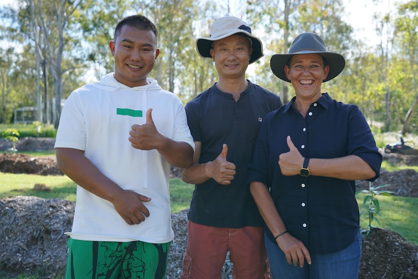 Two chinese men and a woman standing in a sunny orchard, all smiling holding thumbs up. 
