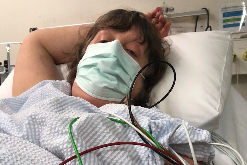 Woman with brown hair sits in hospital bed. She is wearing mask. 