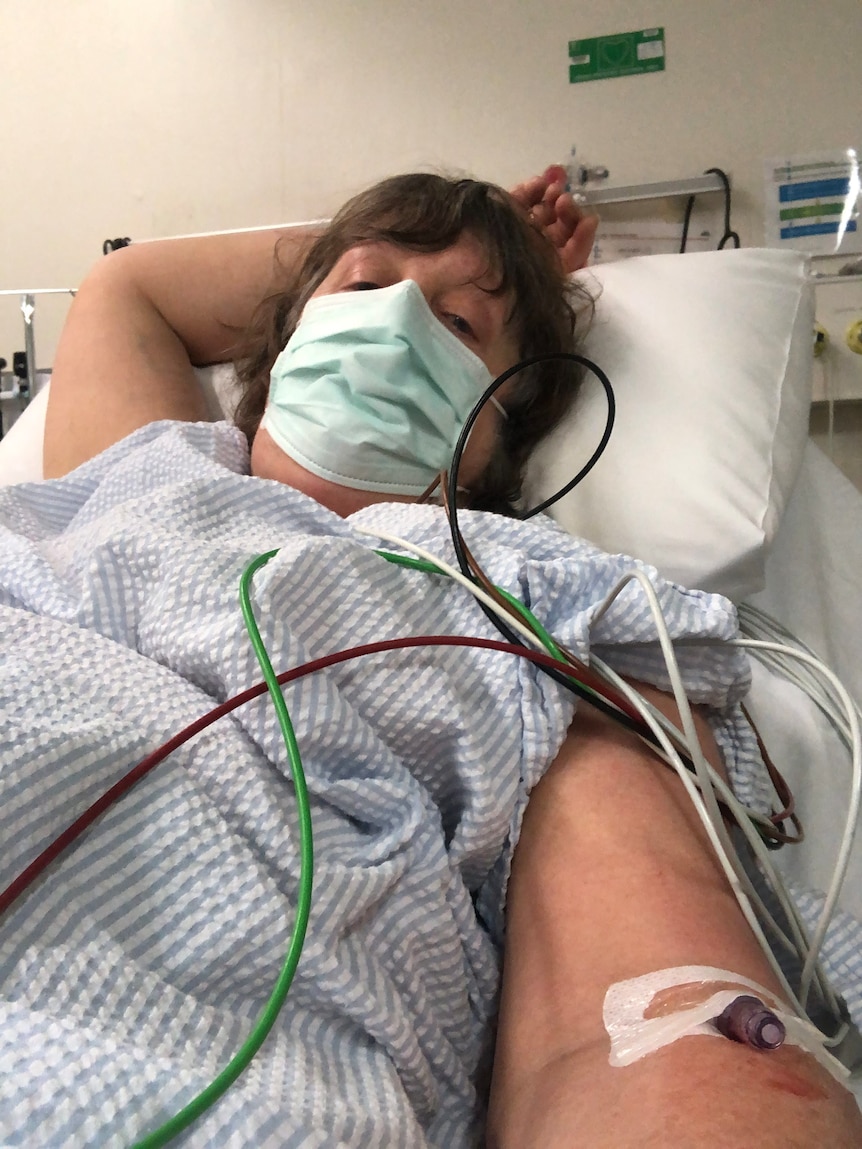 Woman with brown hair sits in hospital bed. She is wearing mask. 