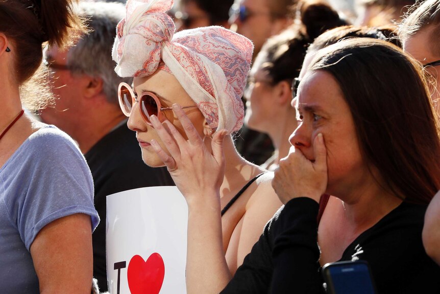A woman adjusts her sunglasses, wiping tears from her eyes and wears a pink pastel scarf on her head.