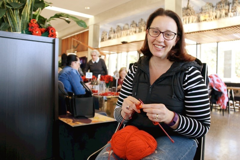 A woman sits on a chair with a ball of red wool to knit decorative poppy flowers.