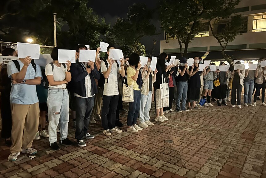 Protesting students hold up blank white papers over their faces as they stand in a line.