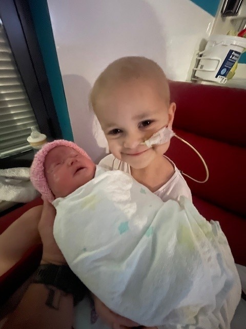 Una niña de 4 años con un tubo en la nariz sonríe mientras sostiene a un bebé recién nacido en una sala de hospital.
