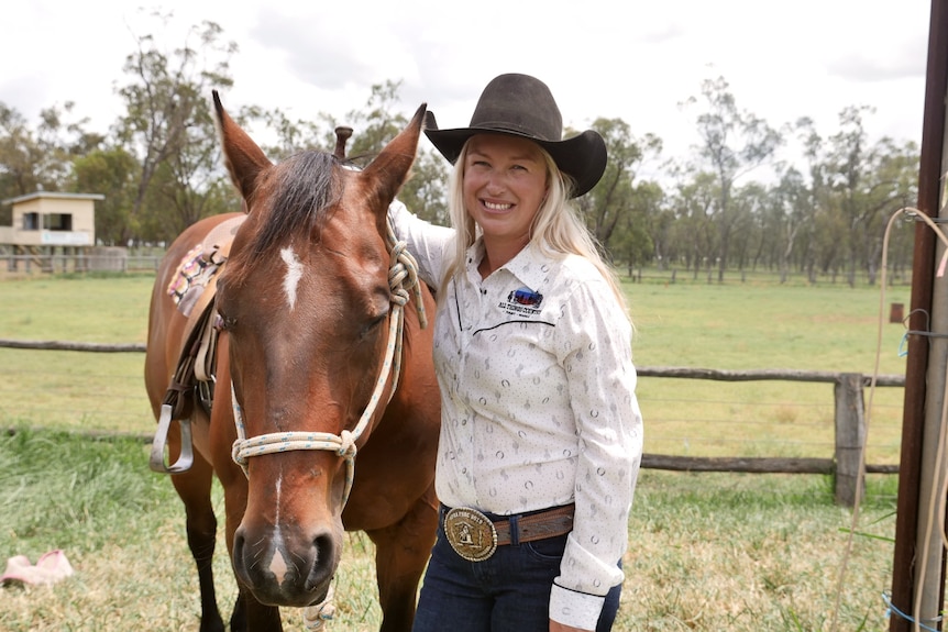 A woman with long blonde hair wearing a black wide brimmed hat stands beside a brown horse and smiles at the camera