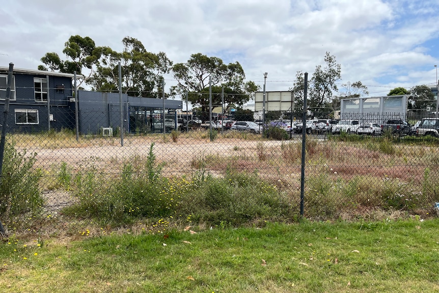 a fence shows a concreted site with overgrown weeds, in the background there is a navy building and a carpark