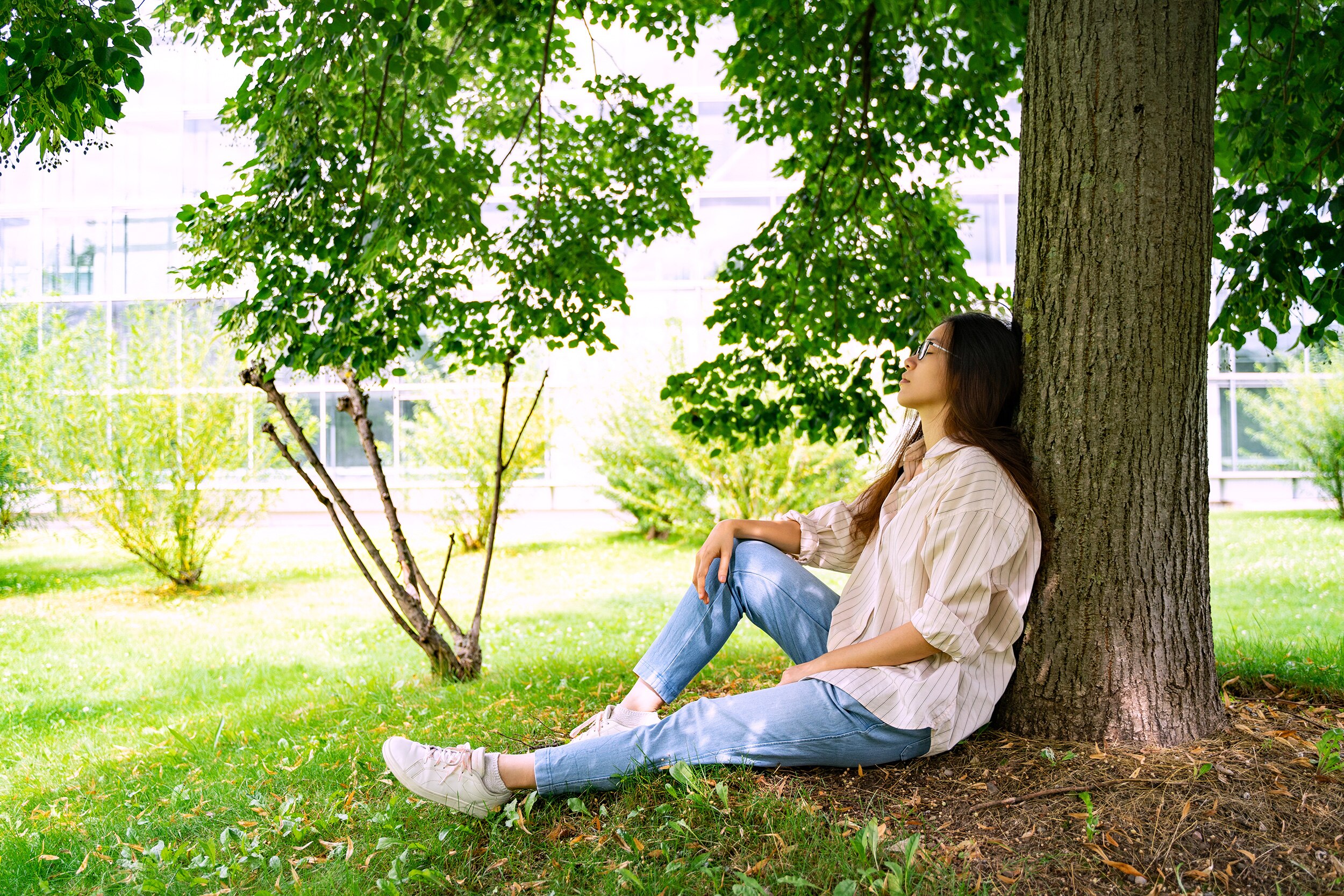 A woman with long brown hair and glasses sits under a tree in the shade, a glass building is in the background.