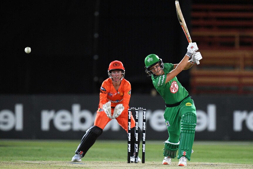 A cricket player wearing green kit hits a ball while a wicketkeeper wearing orange watches on.