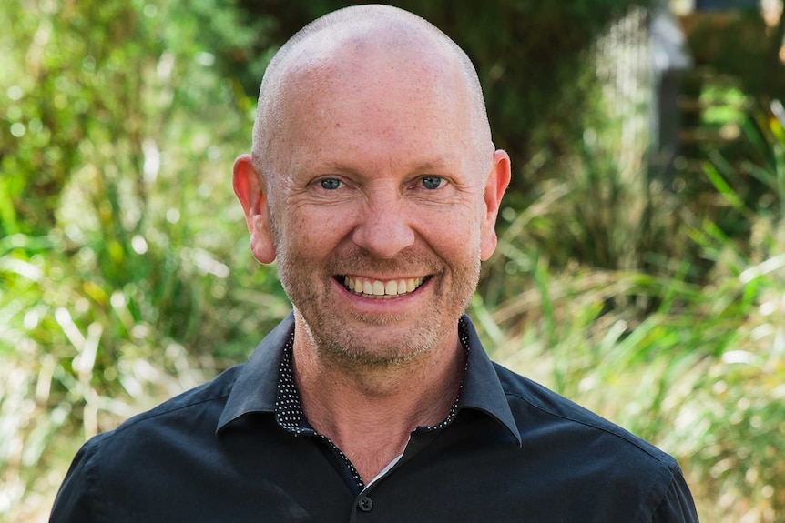 A bald man in a black shirt smiles for the camera. He is standing in a forested area.