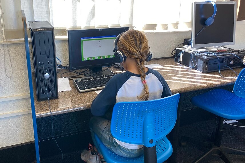 A girl with headphones sitting on a blue chair at a computer doing school work