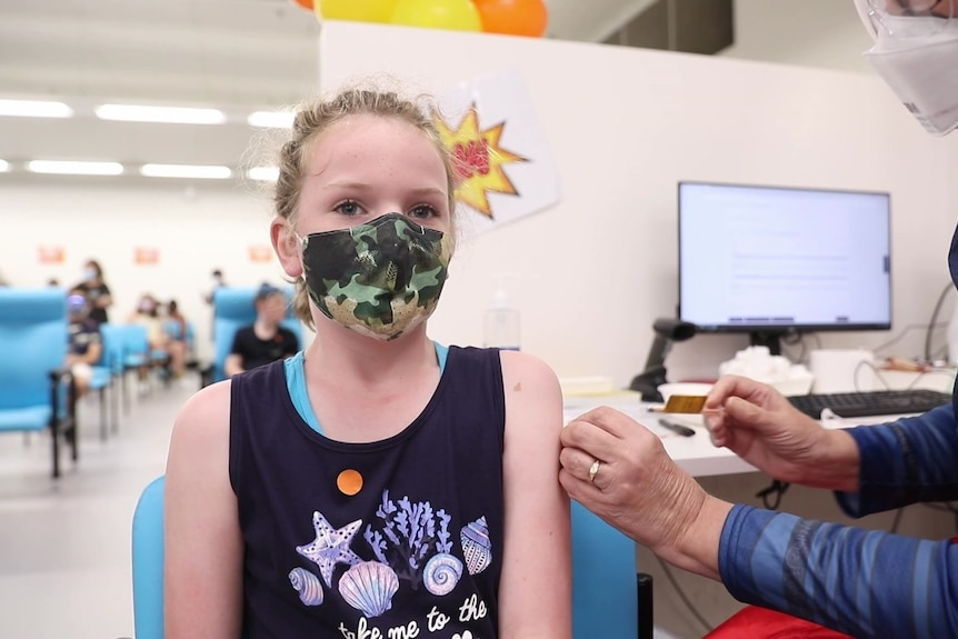 A young girl wearing a singlet and a mask after her vaccine, a nurse holds a small wad  of cotton wool on her upper arm