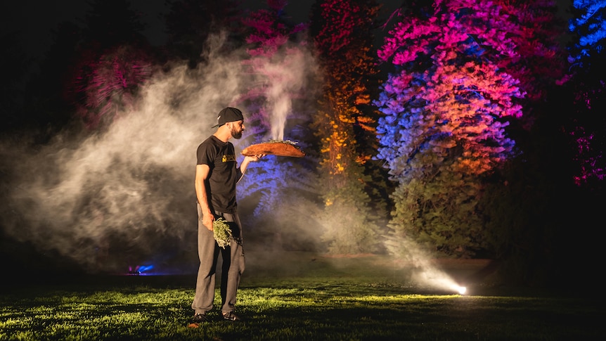 A man performs a welcome to country with smoking wood at night time in front of lit up trees