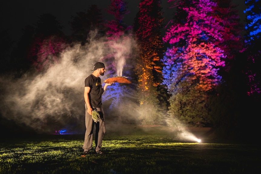 A man performs a welcome to country with smoking wood at night time in front of lit up trees