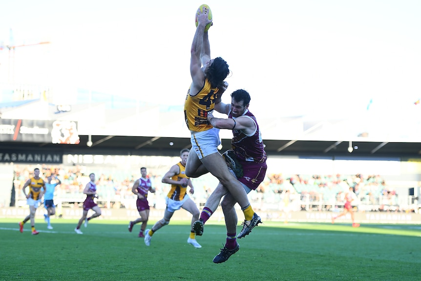 A Hawthorn player leaps high and catches the ball above his head at full stretch as a Brisbane player tackles him.. 