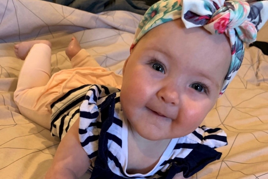 A seven-month old baby lies on her front on a bed, wearing a bright headscarf.