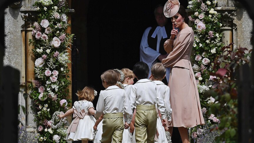 The Duchess of Cmabridge wears a dusty pink gown and matching headpiece and gestures at six children to be quiet entering church