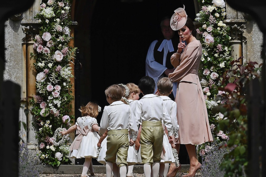 The Duchess of Cmabridge wears a dusty pink gown and matching headpiece and gestures at six children to be quiet entering church